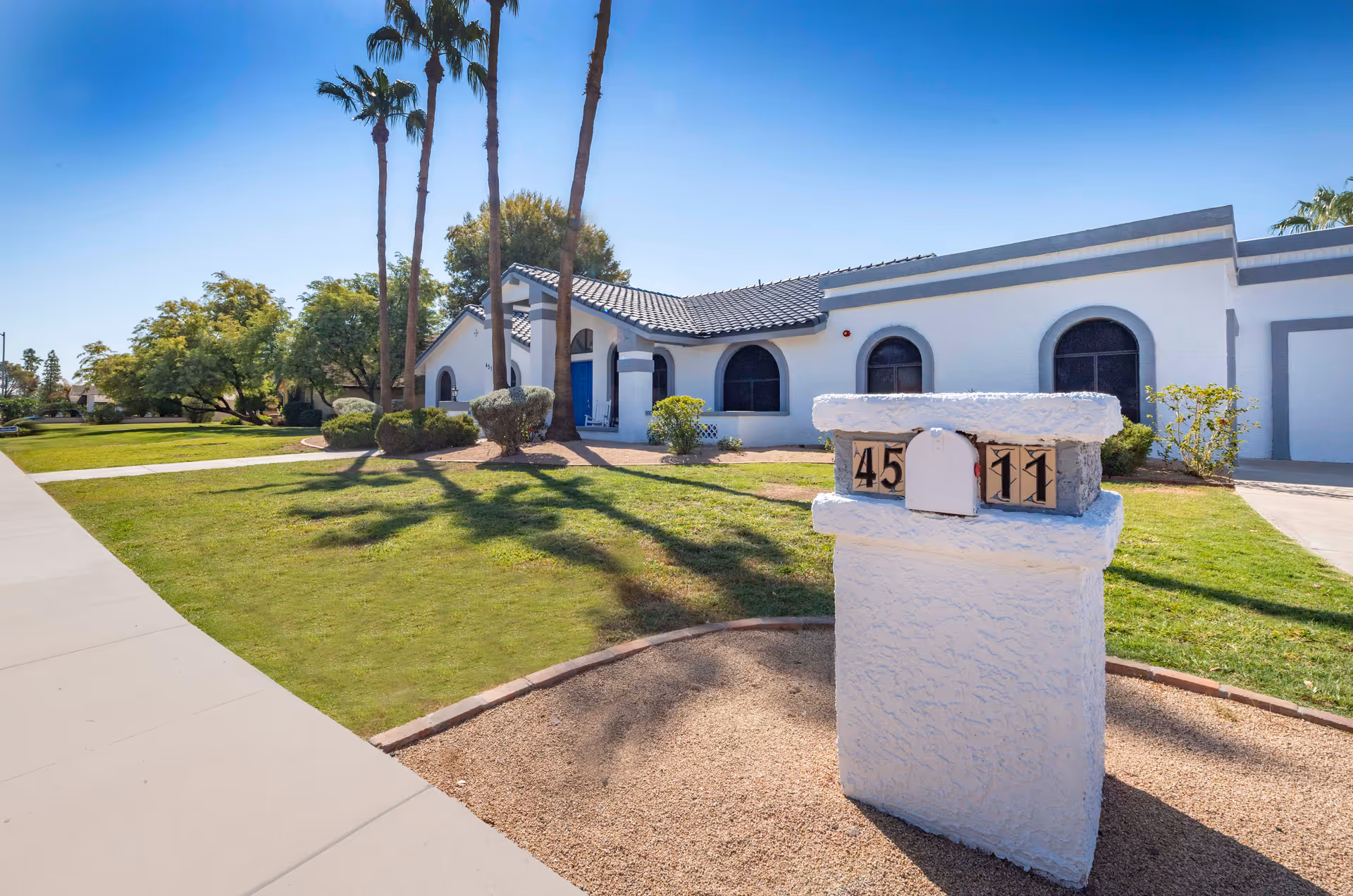Single-story white stucco building with arched windows, palm trees and a white mailbox on a landscaped lawn.