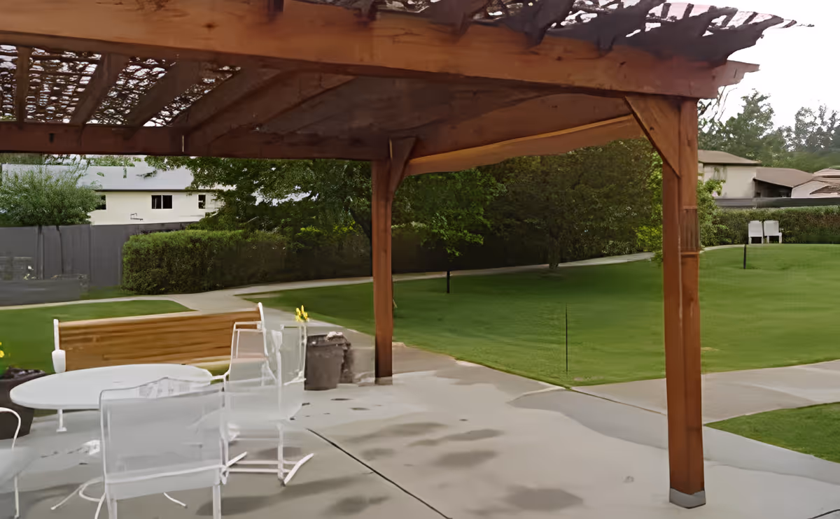 Covered wooden pergola over a patio with white chairs and a table facing a manicured lawn and walkways.