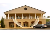 Front exterior view of a two-story beige building with a triangular roof, featuring two symmetrical curved staircases leading to a balcony with white railings. A car is parked in front of the building.