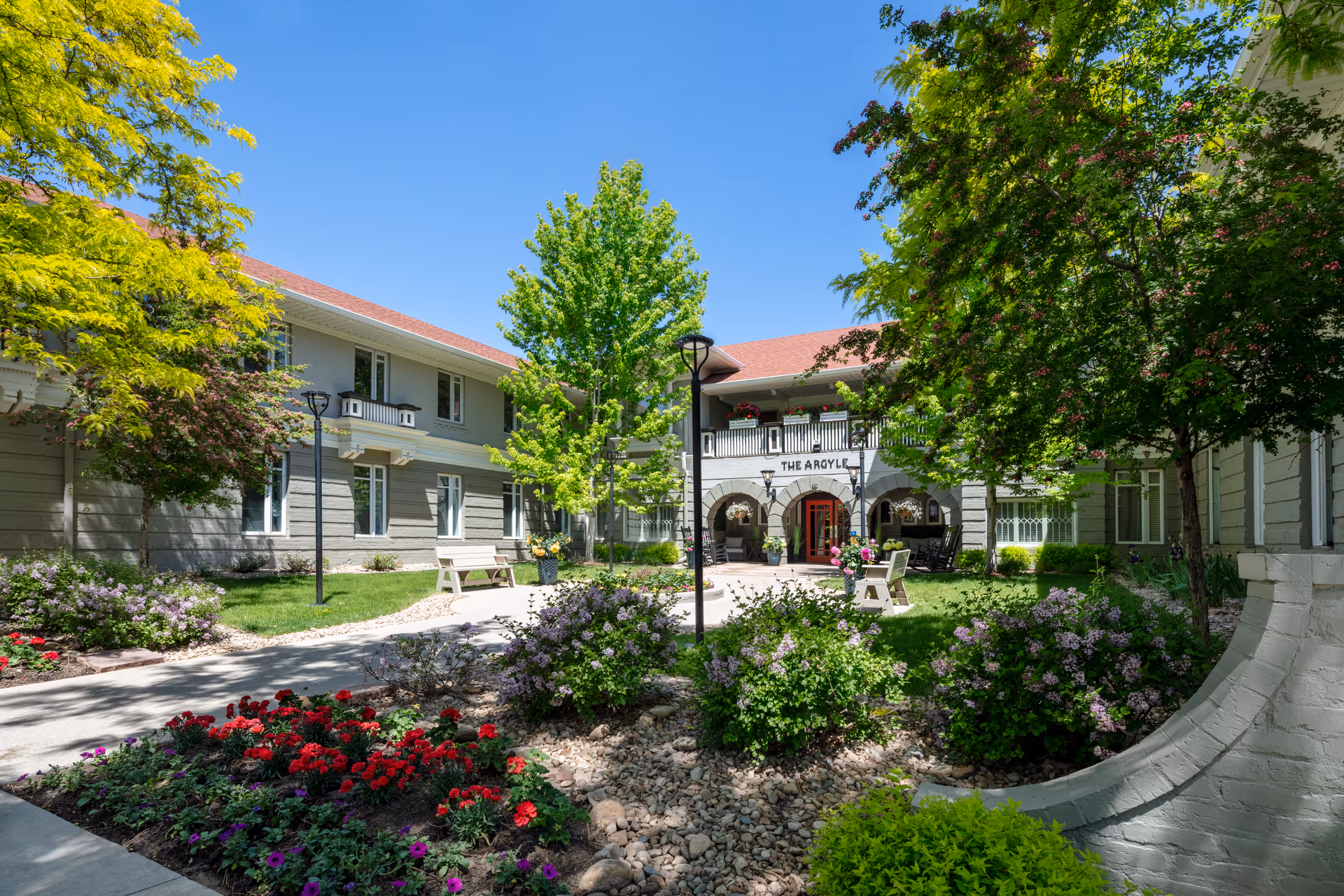 Landscaped courtyard and main entrance of The Argyle building with flowerbeds, benches, and trees under a clear blue sky.