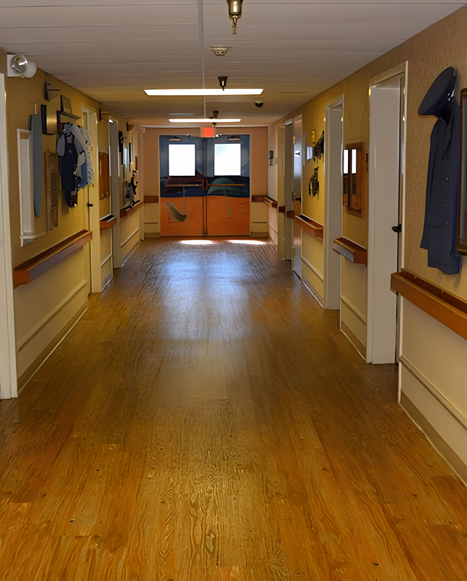 A long hallway with wooden flooring and beige walls, featuring several closed white doors on both sides. The walls are decorated with various framed items and clothing pieces. At the end of the hallway, there are double doors with windows letting in light.