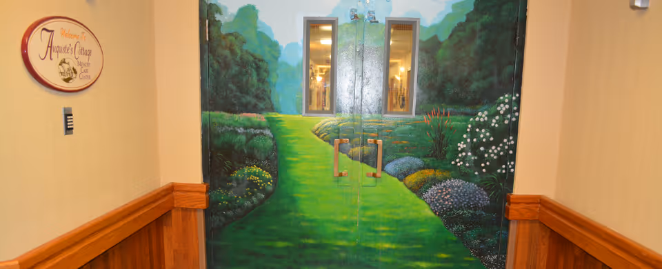Double doors painted with a mural of a green garden path surrounded by bushes and flowers, with two small windows in the doors. To the left of the doors is a sign that reads 'Welcome to Auguste's Cottage Memory Care Center'. The hallway walls are beige with wooden wainscoting.