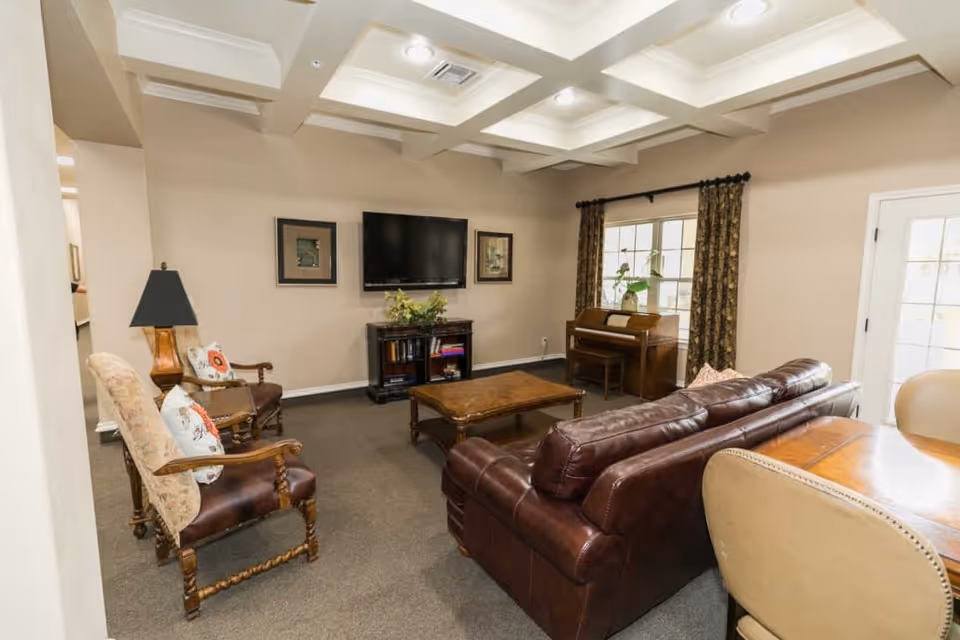 A cozy living room with a brown leather sofa, two upholstered armchairs with wooden frames, a wooden coffee table, a flat-screen TV mounted on the wall above a small cabinet, a piano near a window with patterned curtains, and a round wooden table with beige chairs. The room has beige walls and a ceiling with recessed lighting.
