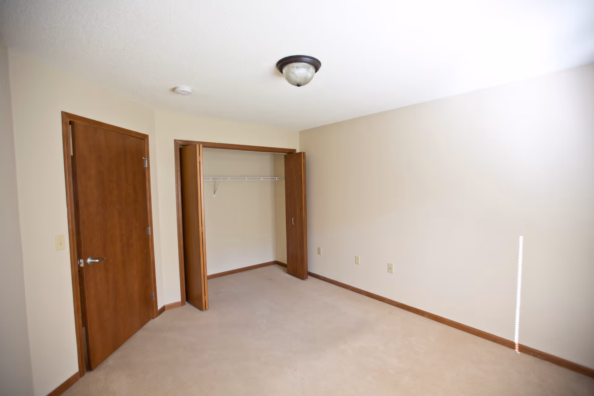 Empty carpeted bedroom with an open closet, wooden doors, beige walls and a ceiling light.