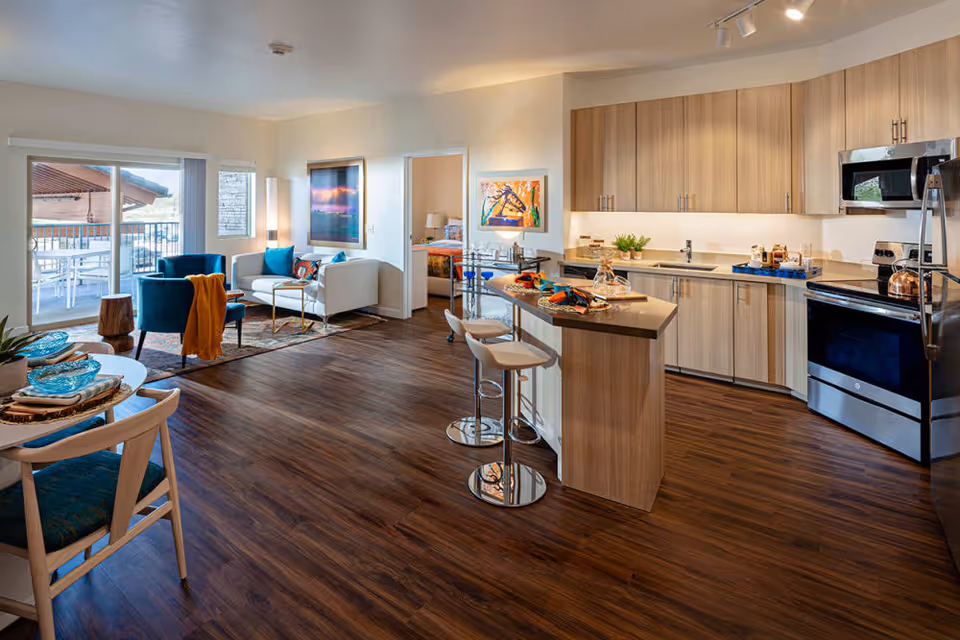 A modern open-concept living space in a senior living facility featuring a kitchen with light wood cabinets, stainless steel appliances, and a breakfast bar with two stools. Adjacent to the kitchen is a living area with a white sofa, two blue armchairs, a small coffee table, and a sliding glass door leading to an outdoor patio with a table and chairs. A bedroom is visible through an open door, and the room has wooden flooring and bright, natural lighting.