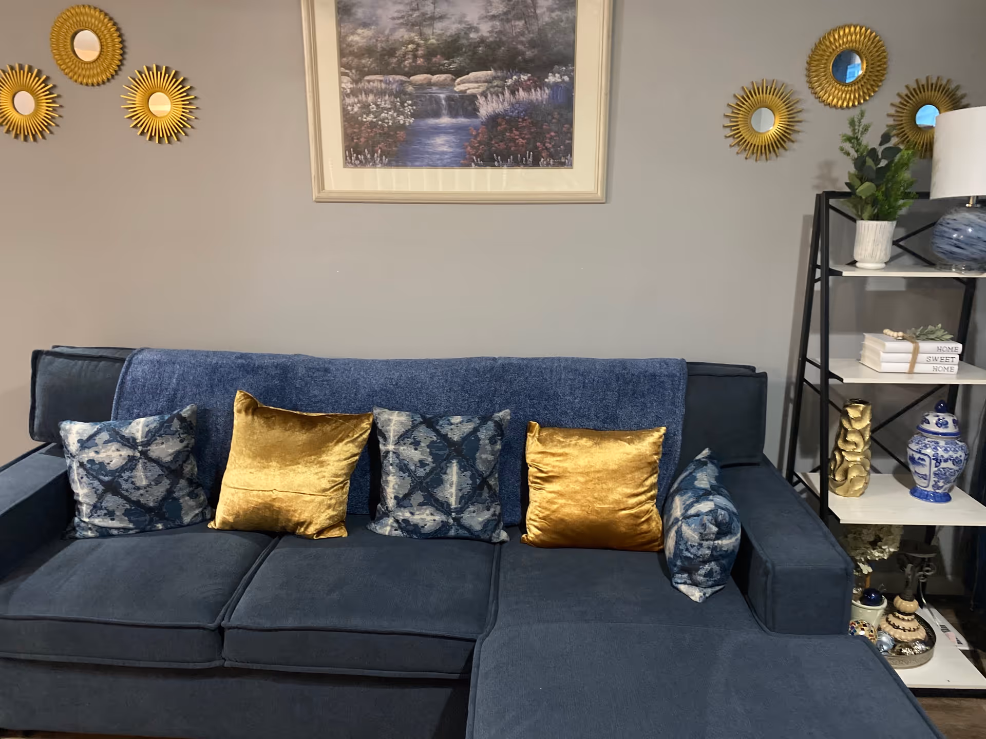 A cozy living room area featuring a dark blue sectional sofa adorned with four decorative pillows in gold and blue patterns. Above the sofa hangs a framed painting of a serene waterfall scene. The wall is decorated with five small round mirrors with sunburst gold frames. To the right of the sofa is a black metal and white shelf unit holding various decorative items including plants, books, and blue and white ceramic pieces.