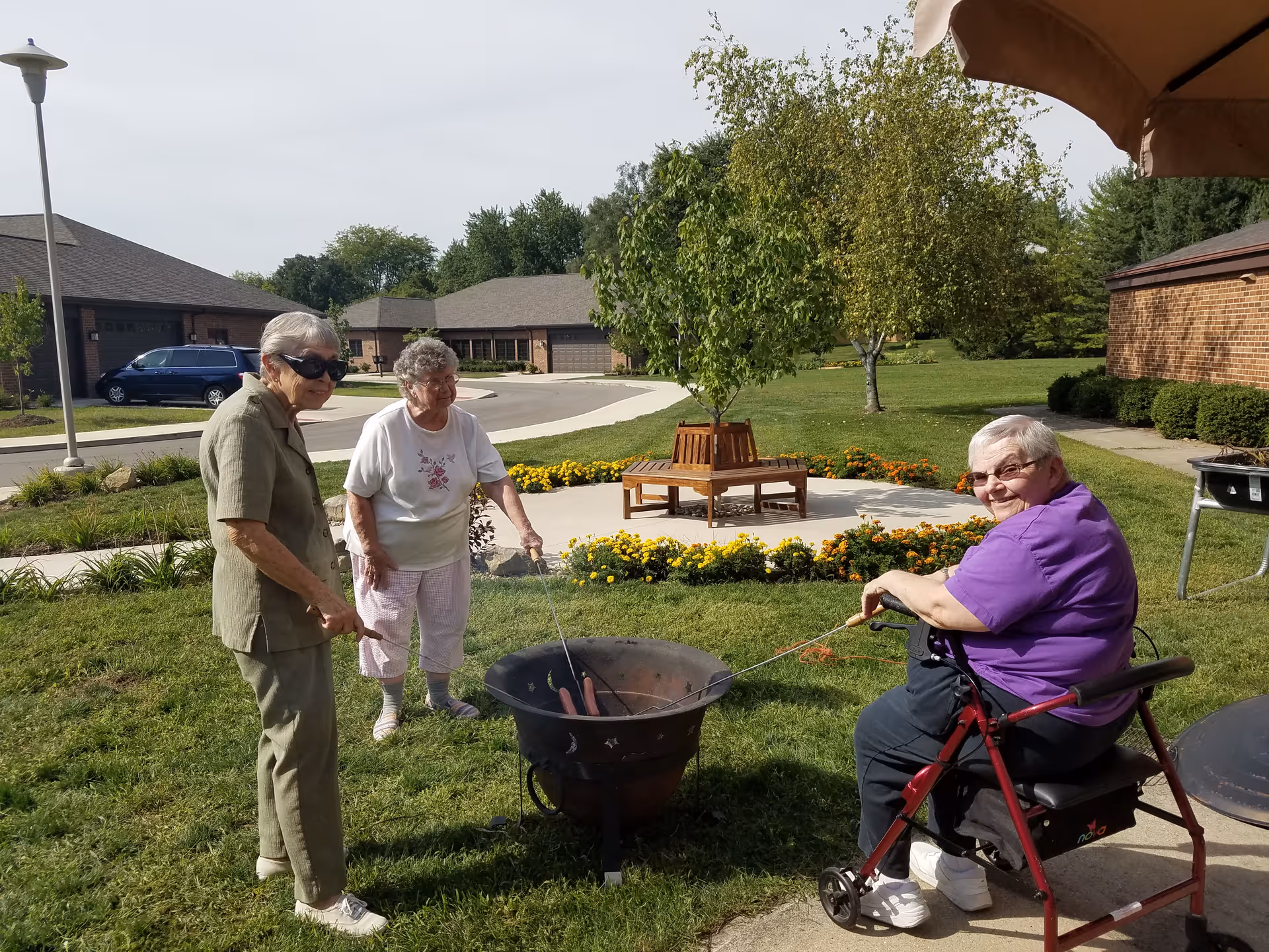 Three elderly women outdoors at a senior living facility. Two women are standing and roasting hot dogs over a fire pit, while the third woman, seated on a walker, is also roasting hot dogs. The background shows a well-maintained garden with flowers, trees, and residential buildings under a clear sky.