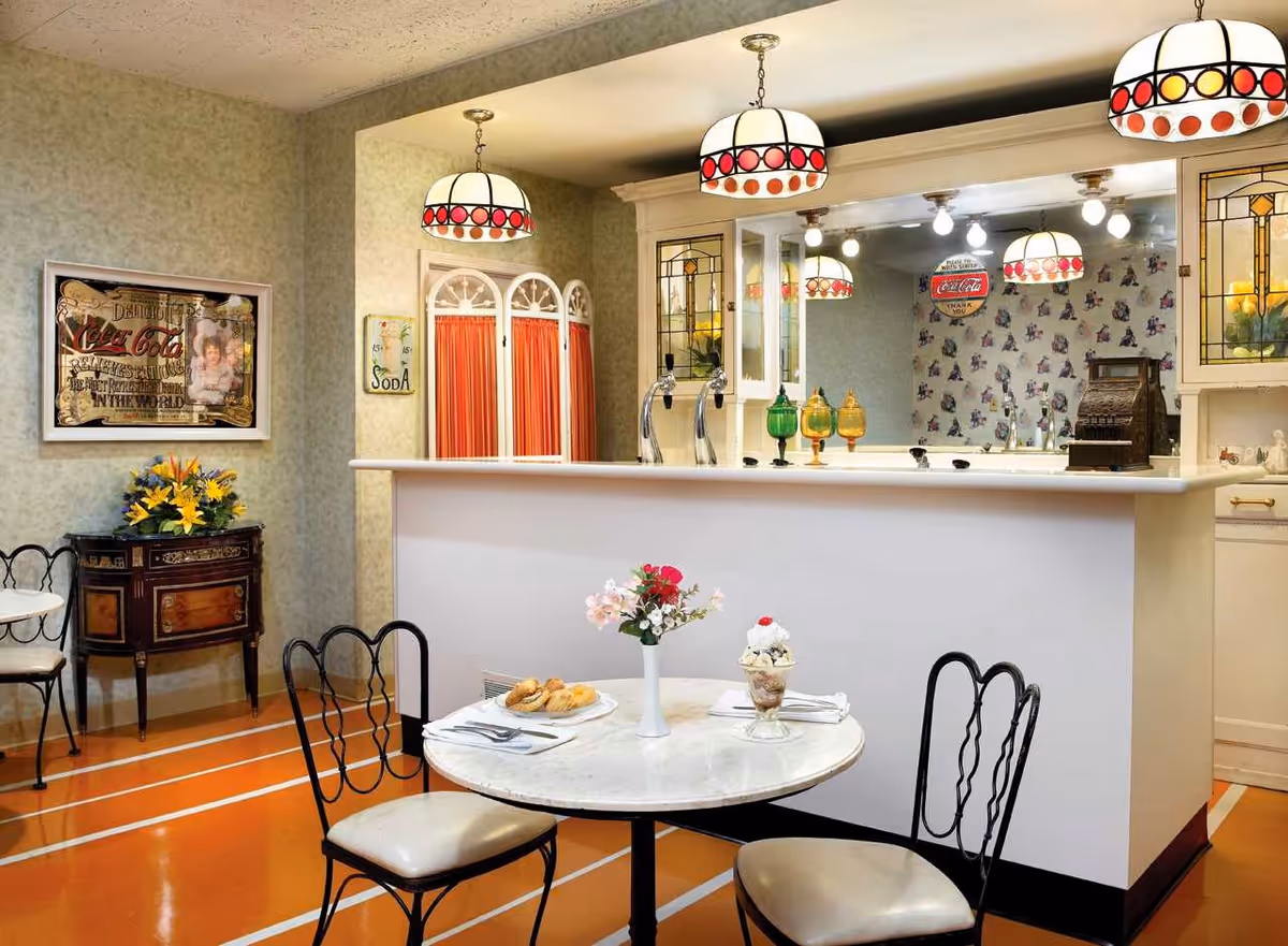 A cozy dining area with a small round table set with pastries, a dessert, and a vase of flowers. The table is surrounded by three chairs with decorative backs. Behind the table is a white counter with vintage-style soda taps and colorful glass containers. Above the counter hang three pendant lights with stained glass shades. The walls are decorated with vintage Coca-Cola signs and floral wallpaper, and there is a small wooden cabinet with a flower arrangement on the left side.