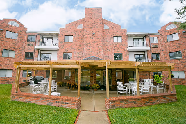 Brick multi-story building with a covered wooden pergola patio, white outdoor chairs, and a grassy lawn in front.