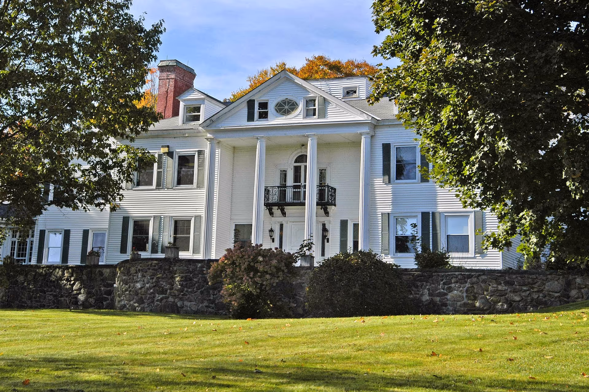 Large white colonial-style building with tall white columns at the entrance, green shutters on the windows, a stone wall at the base, surrounded by trees and a well-maintained lawn under a blue sky.