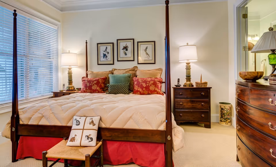 A cozy bedroom with a wooden four-poster bed adorned with a beige quilt and multiple decorative pillows in red, green, and patterned designs. On either side of the bed are wooden nightstands with matching lamps. Above the bed are three framed bird illustrations. To the right, there is a wooden dresser with a lamp and decorative items. A window with white blinds is on the left side of the room.
