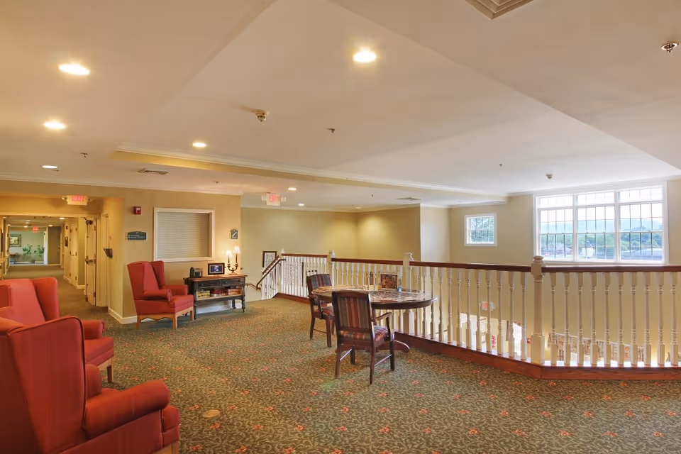 A spacious, well-lit interior common area in a senior living facility with red upholstered armchairs, a small table with chairs, carpeted floor, and large windows letting in natural light. The area overlooks a lower floor with a white railing and has a hallway leading to other rooms.