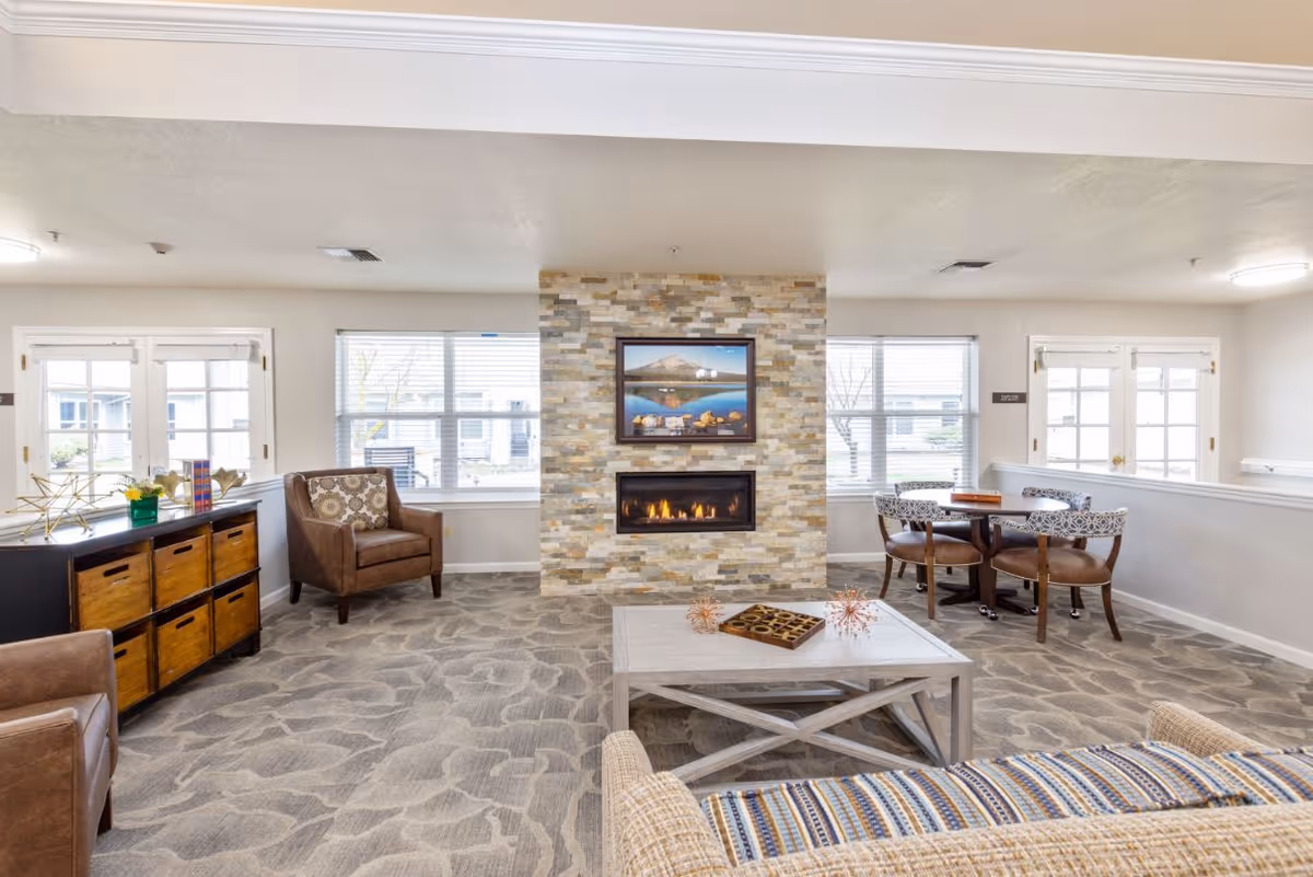 A bright and spacious living room area in a senior living facility featuring a modern stone fireplace with a framed landscape picture above it. The room has large windows letting in natural light, a patterned carpet, a wooden coffee table with decorative items, comfortable armchairs, a small round table with four chairs, and a wooden storage unit with baskets.