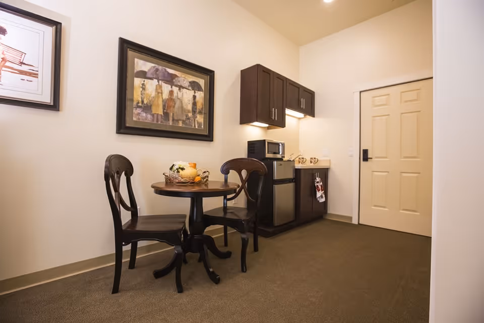 Small interior dining nook with a round table and two chairs beside a compact kitchenette and a closed door.