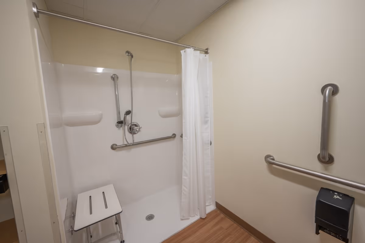 Accessible shower stall with grab bars, a handheld showerhead, folding shower seat and white curtain in a beige-walled bathroom.