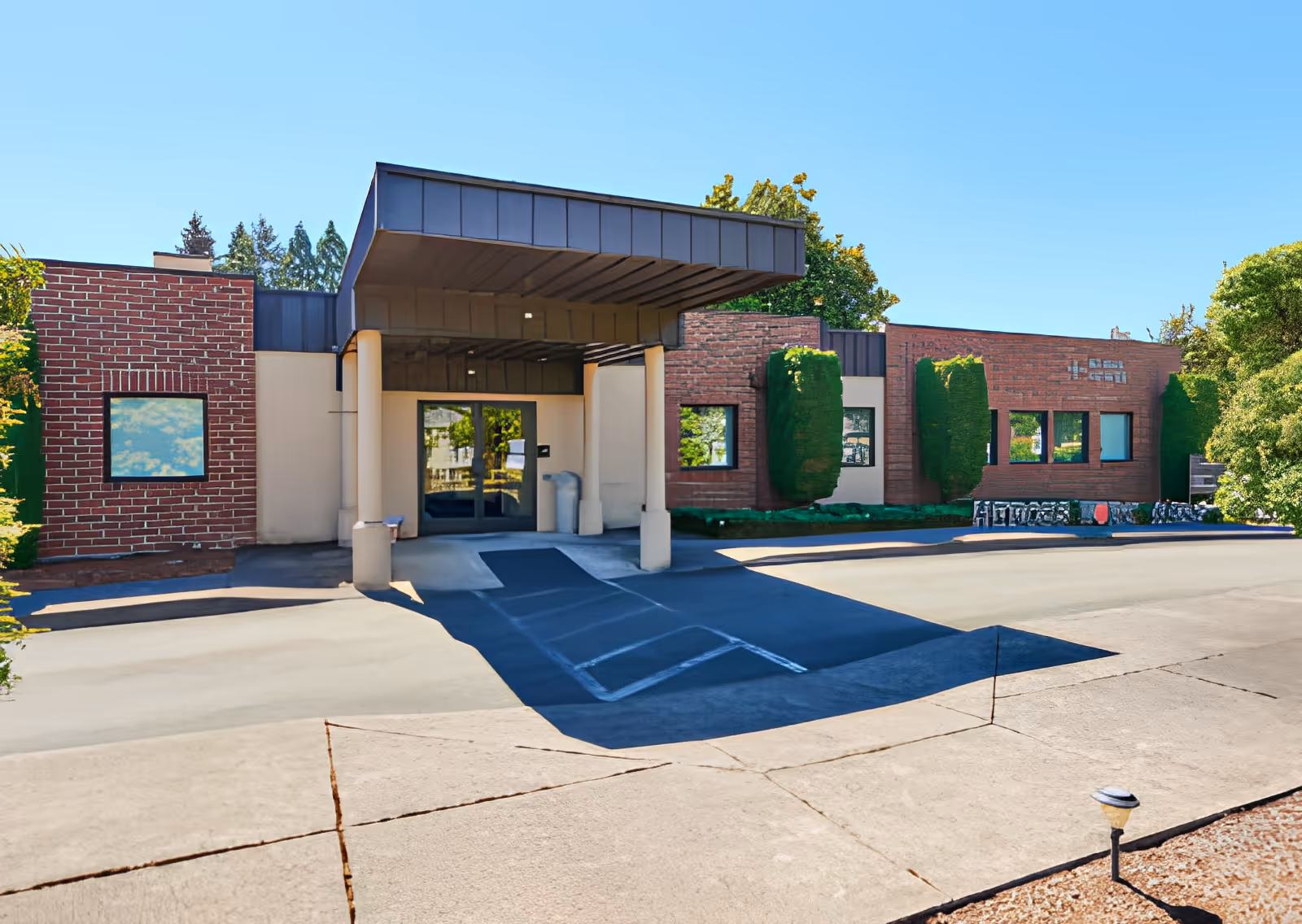 Exterior view of Elliott Residence showing the main entrance with a covered drop-off area, brick and beige walls, windows, and surrounding greenery under a clear blue sky.