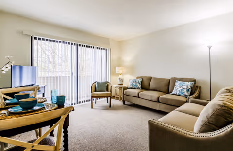 Bright living room with sofas, an armchair, a small dining table, and sliding glass doors leading to a balcony.