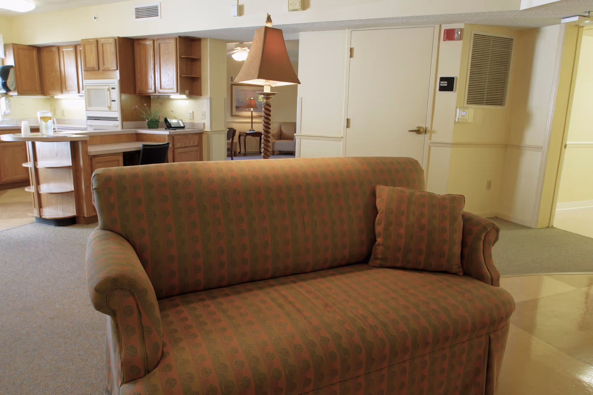 Interior view of a senior living facility showing a patterned sofa with a matching cushion in the foreground. Behind the sofa, there is a wooden kitchen area with cabinets, a microwave, and a countertop. A lamp with a twisted base and a lampshade is positioned near the sofa. The background also includes a hallway with doors and a small seating area with chairs and a table.