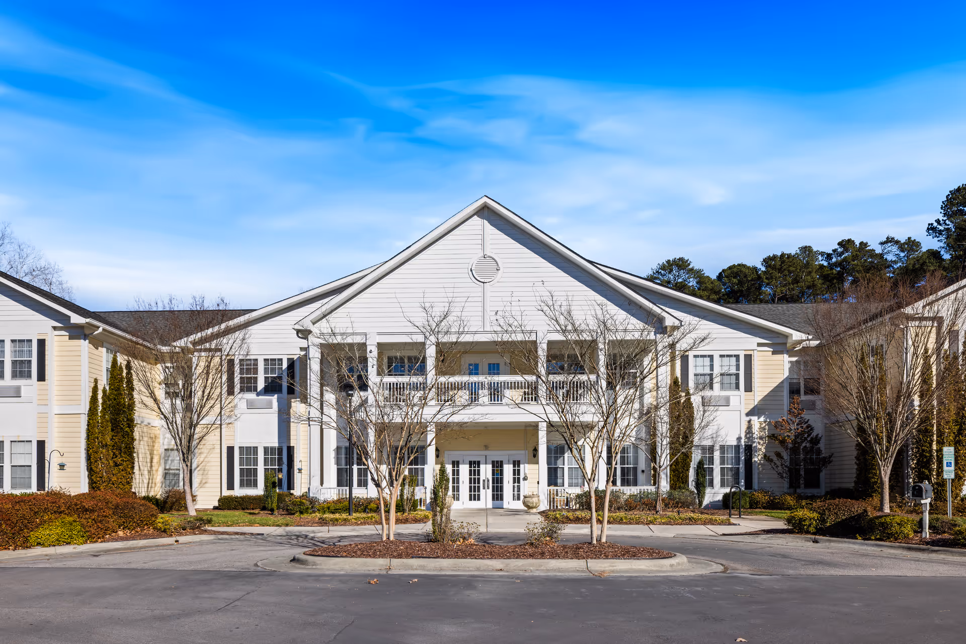 Front exterior of a two-story white senior living building with columns, a covered entrance, balcony, and landscaped driveway.