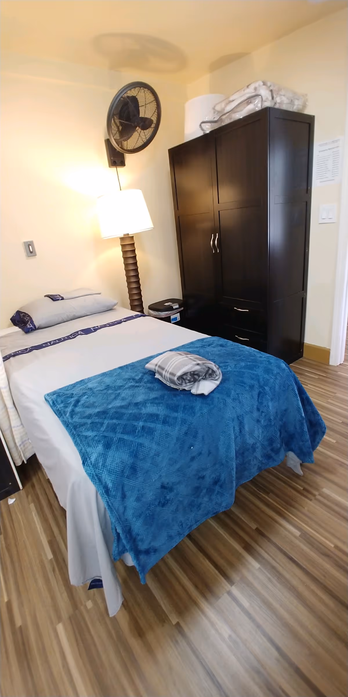 A small bedroom with a single bed covered with white sheets and a blue blanket. A folded gray and white blanket is placed on top of the bed. Next to the bed is a tall floor lamp and a wall-mounted fan. There is a dark wooden wardrobe with some folded blankets on top. The floor has a wood-like finish.