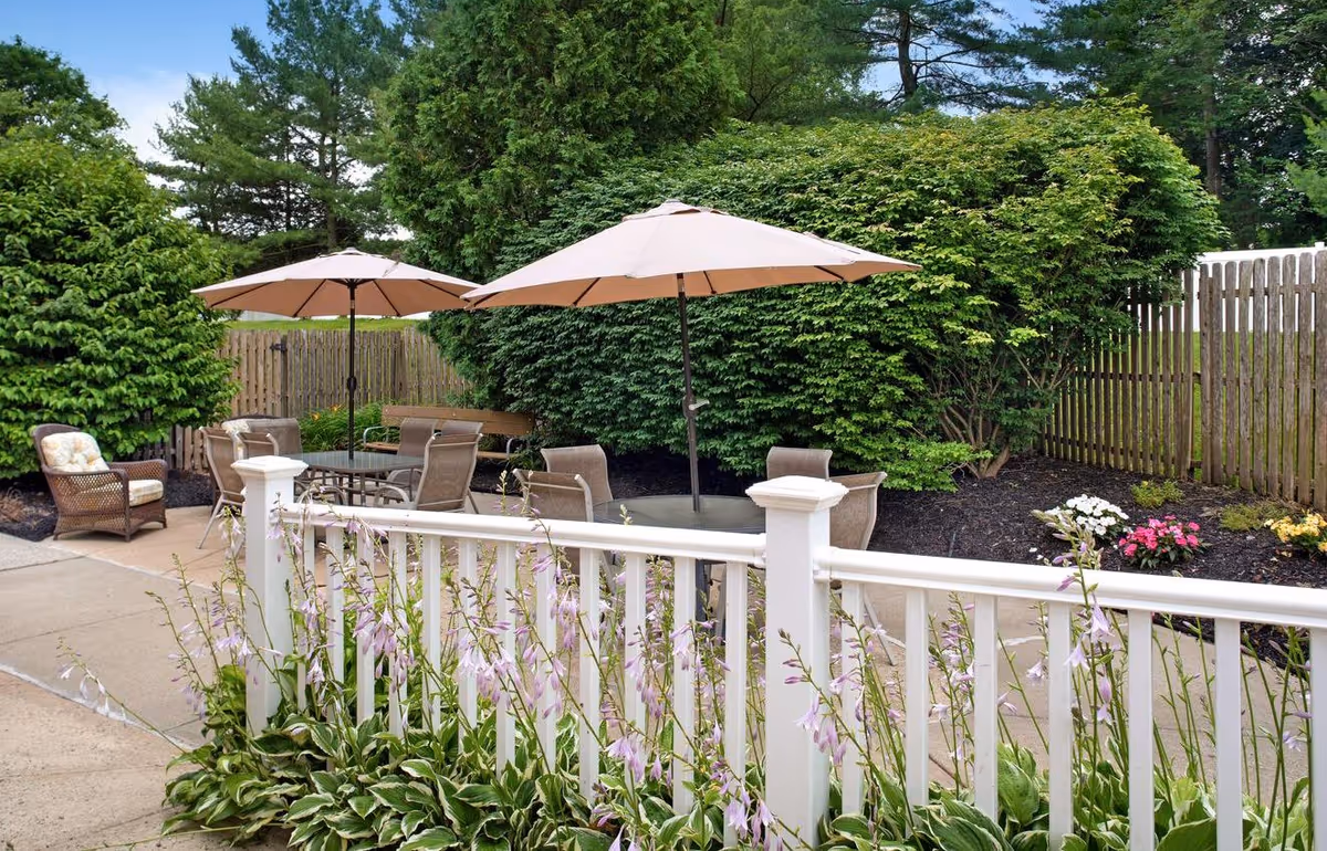 A fenced outdoor patio featuring tables with umbrellas, chairs, a white railing, and surrounding shrubs and flowers.