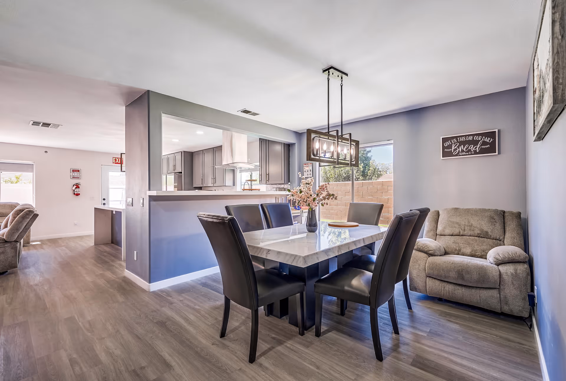 A modern dining area with a rectangular marble table surrounded by six black leather chairs. A vase with pink flowers is placed on the table. To the right, there is a comfortable gray armchair against a light purple wall with a framed sign that reads 'Give us this day our daily bread Matthew 6:11'. The dining area is adjacent to a kitchen with gray cabinets and a stainless steel range hood. The floor is covered with light wood laminate, and a large window lets in natural light.