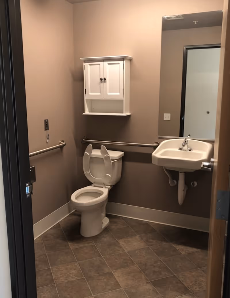 A small bathroom with beige walls and tiled floor. It features a white toilet with the seat and lid raised, a white wall-mounted sink with a faucet, a large rectangular mirror above the sink, and a white wall cabinet with two doors above the toilet. There are metal grab bars on the walls beside and behind the toilet for accessibility.