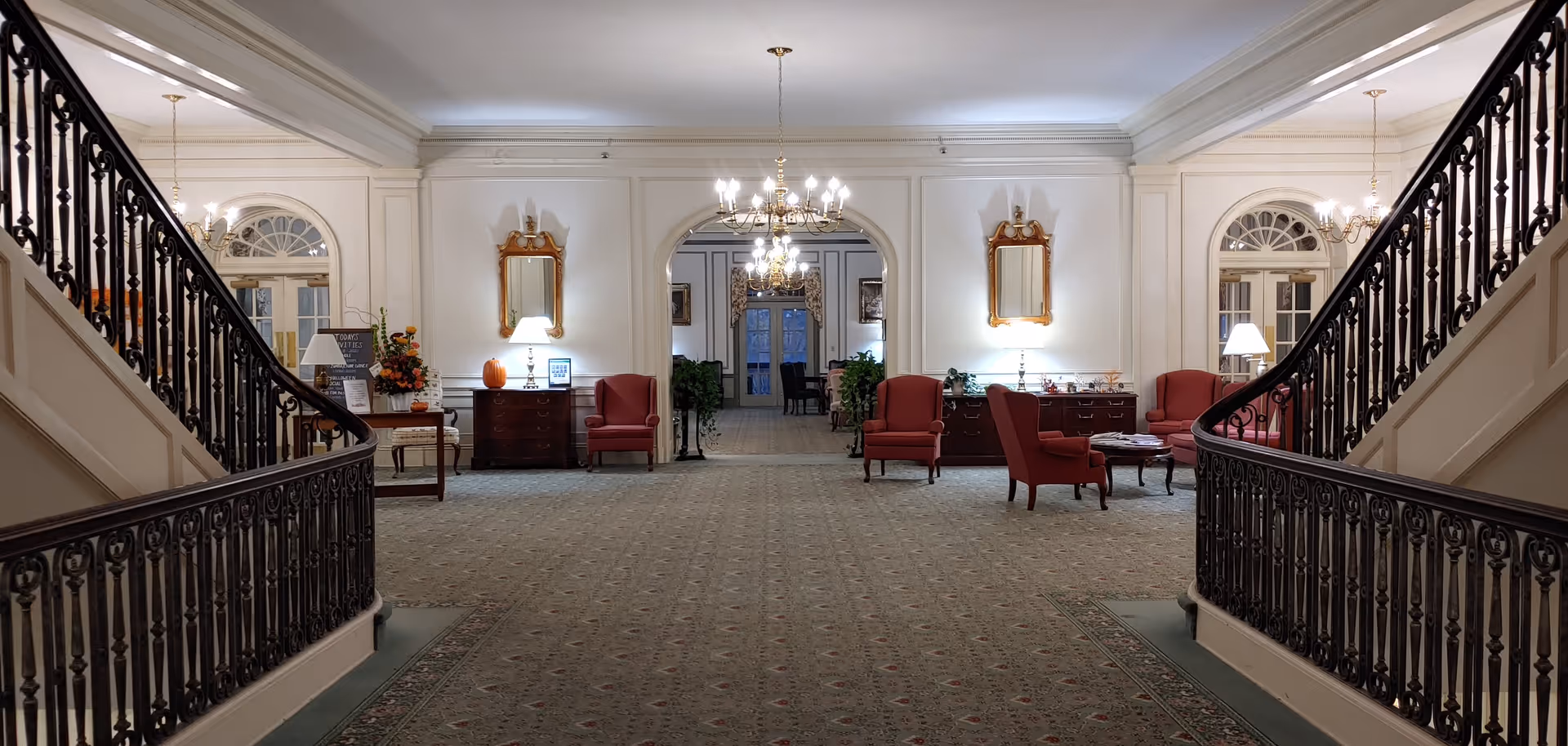 Spacious and elegant interior of a retirement facility lobby with two symmetrical staircases featuring ornate black railings. The room has a patterned carpet, white walls with decorative molding, and two chandeliers hanging from the ceiling. There are red upholstered chairs and wooden tables with lamps and decorative items along the walls. Two large mirrors hang above the tables, and an arched doorway leads to another room in the background.