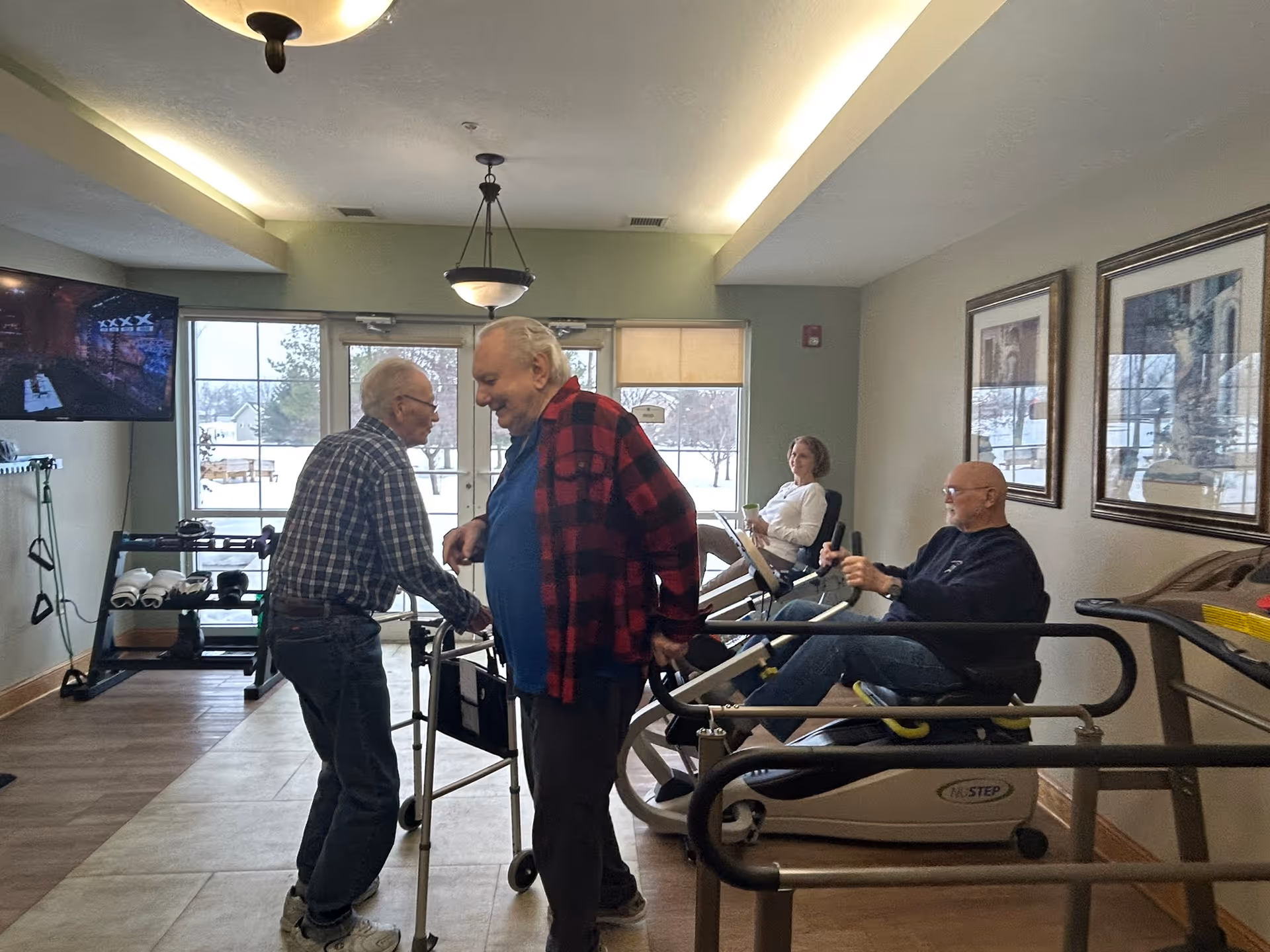 A group of elderly people in a fitness room at Summit Pointe. Two men are standing and interacting near a walker, while a man and a woman are seated on exercise machines in the background. The room has large windows showing a snowy outdoor scene, framed pictures on the wall, and a television mounted on the left side.