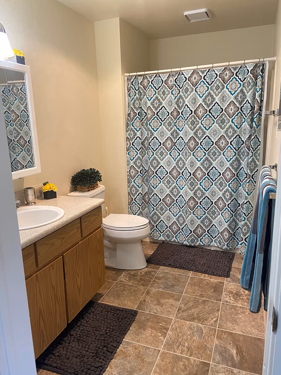 A clean bathroom with a wooden vanity cabinet, a white sink, a toilet, and a shower with a patterned blue, gray, and white curtain. There are two dark brown bath mats on the tiled floor and two blue towels hanging on a towel rack on the right wall. A small plant and a decorative item with yellow flowers are placed on the vanity and toilet tank.
