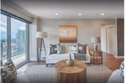Bright modern living room with a white sofa, round wooden coffee table, floor lamp and large sliding glass doors opening to a balcony with city views.