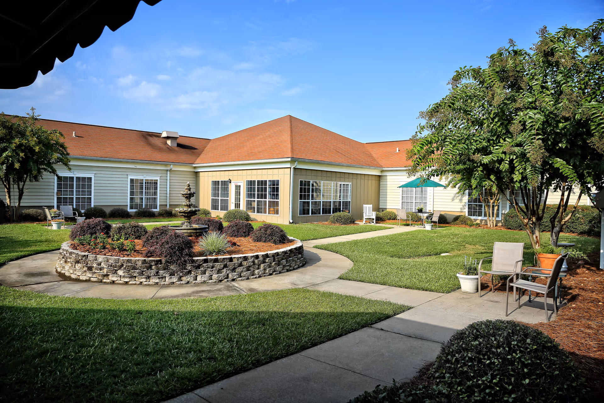 Sunlit courtyard with a circular raised flower bed and fountain surrounded by walkways in front of a single-story senior care building.