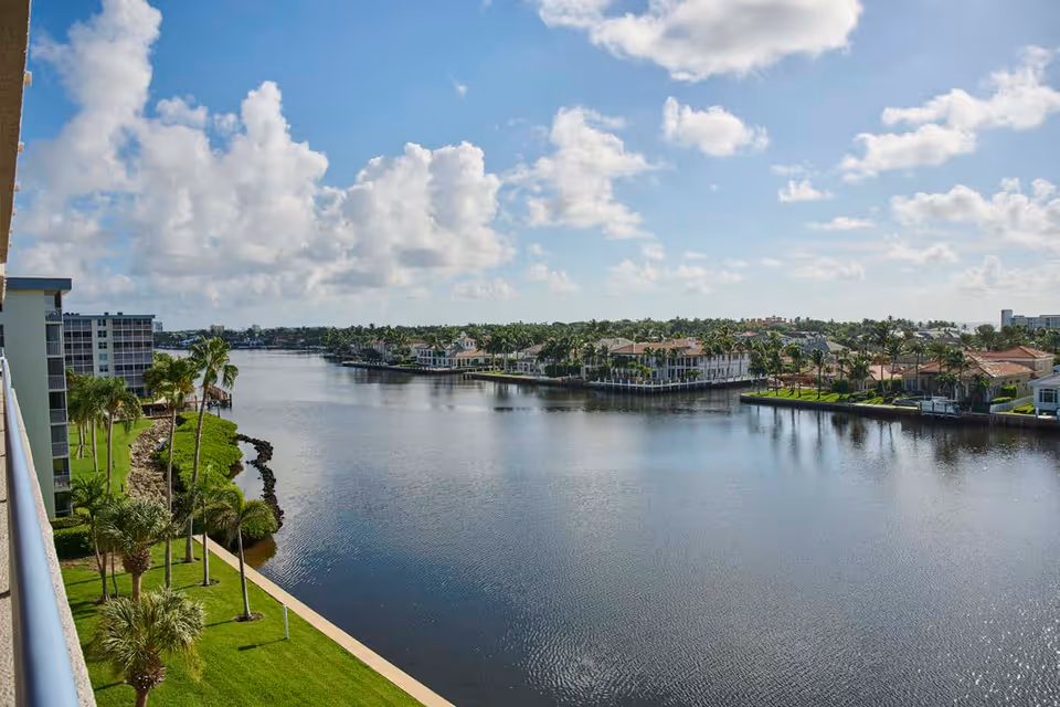 View of a calm waterfront lined with palm trees and residential buildings under a partly cloudy sky.