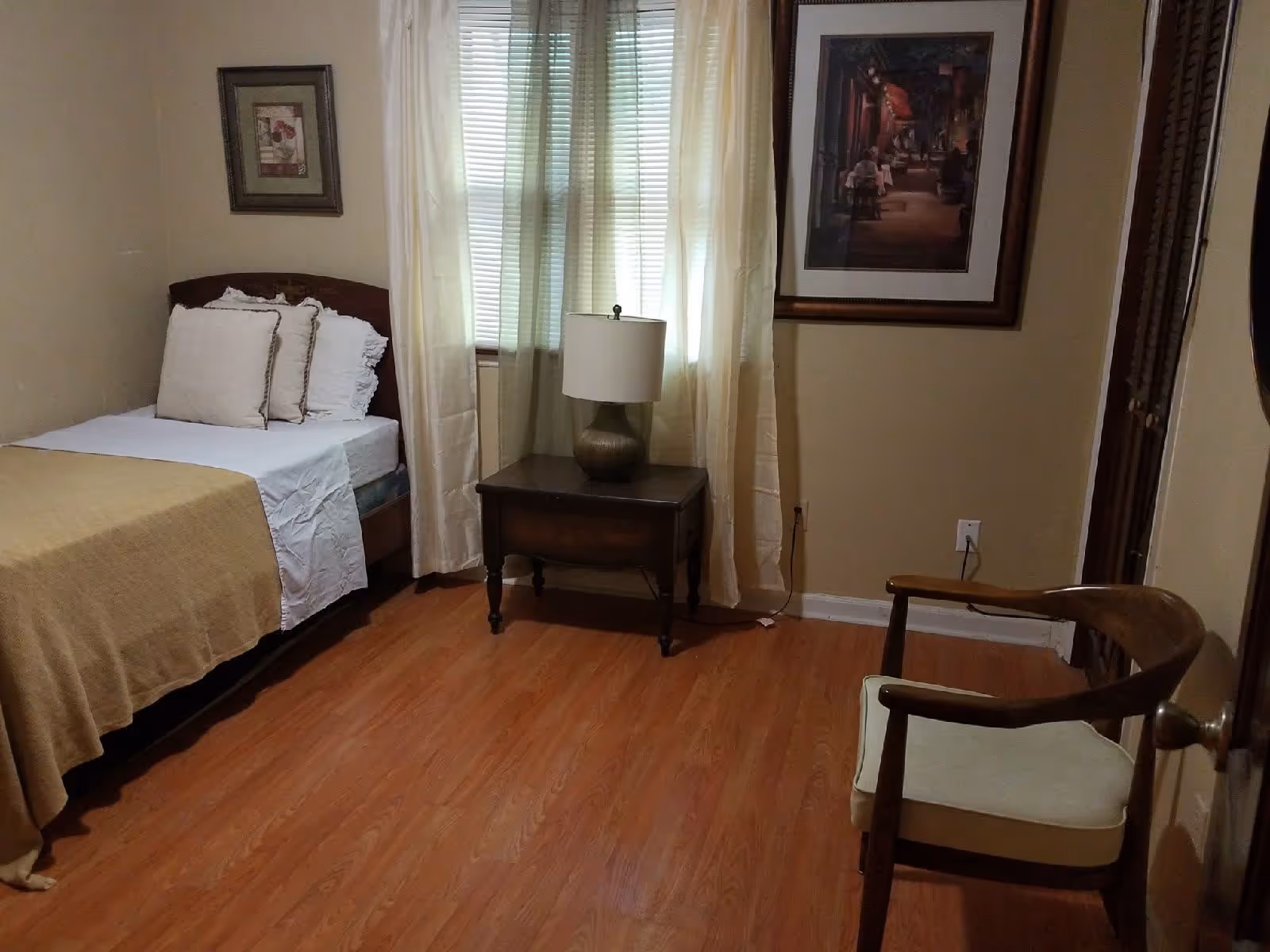 A small bedroom with a single bed covered with a beige blanket and white pillows. Next to the bed is a wooden nightstand with a lamp. The room has wooden flooring, a window with white curtains, a framed picture on the wall above the nightstand, and a wooden chair with a cushioned seat near the door.