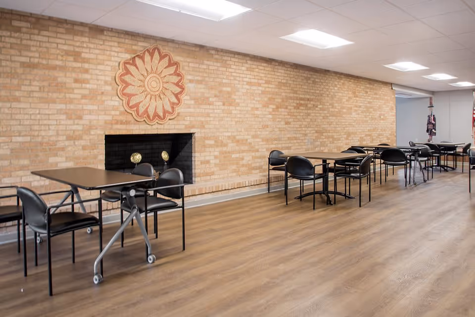 A spacious room with multiple tables and black chairs arranged neatly on a wooden floor. The room features a brick wall with a decorative circular wall hanging above a non-functional fireplace. The ceiling has several rectangular fluorescent lights, and there is an open doorway visible at the far end of the room.