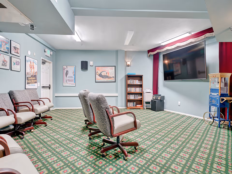 A cozy common room with patterned green carpet and light blue walls. The room features several cushioned chairs with wooden armrests arranged facing a large flat-screen TV mounted on the wall. There is a popcorn machine on the right side and a bookshelf filled with DVDs or books against the back wall. Vintage movie posters decorate the walls, and the room is softly lit with ceiling and wall lights.