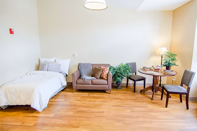 Bright studio-style bedroom with a single bed, a loveseat, a small round dining table with two chairs, and potted plants on hardwood floors.