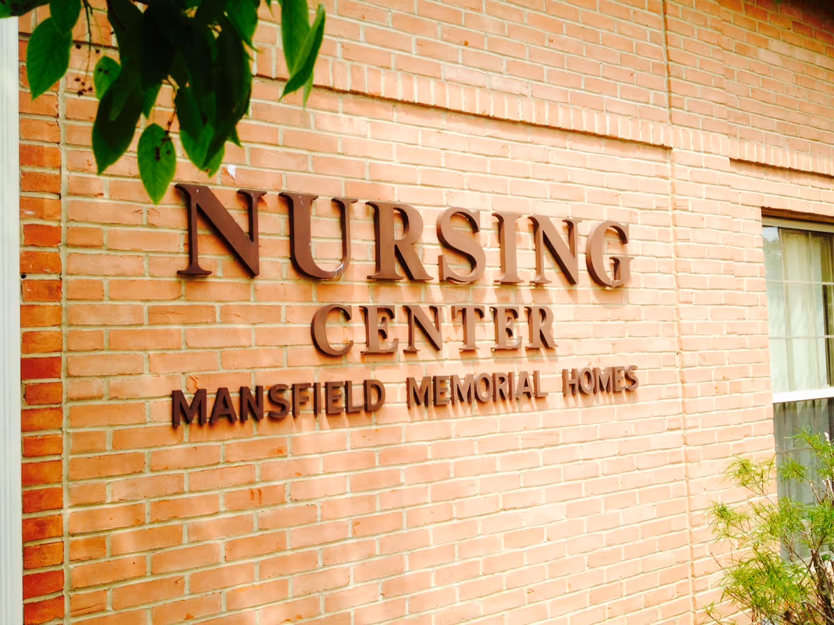 Brick wall with raised brown letters spelling out 'NURSING CENTER MANSFIELD MEMORIAL HOMES' with some green leaves and a window partially visible on the right side.