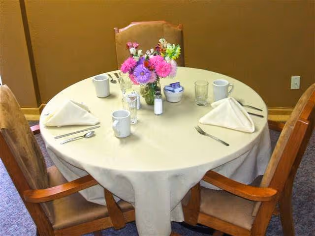 Round dining table set for four with napkins, mugs, utensils and a vase of flowers.