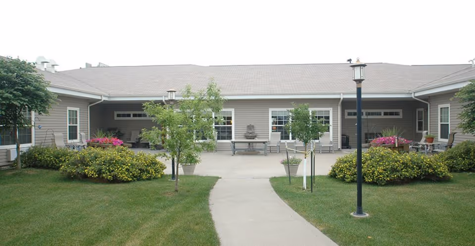 Walkway leading into a landscaped courtyard in front of a single-story assisted living building with benches, planters, and lamp posts.
