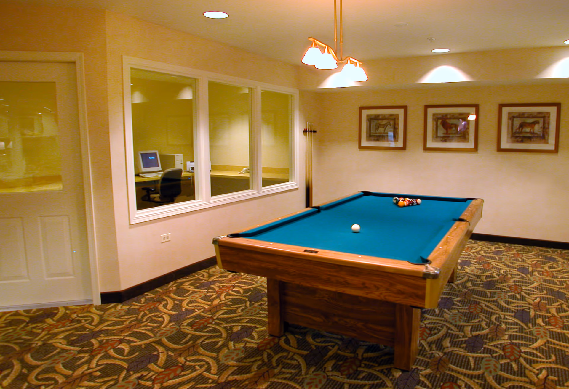 Indoor recreation room with a pool table, overhead lights, framed wall art and a computer workstation visible through interior windows.