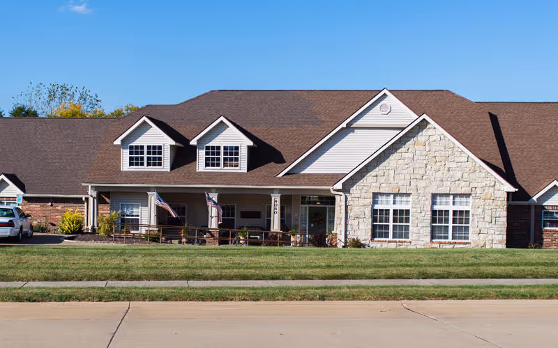 Exterior view of a single-story senior living facility building with a brown roof, stone and brick facade, multiple windows, a covered porch with two American flags, and a well-maintained lawn in front.