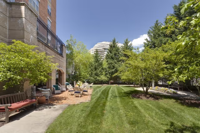 Outdoor garden area at Brighton Gardens of Friendship Heights featuring a well-maintained lawn, several trees, a brick patio with white metal chairs and tables, and a wooden bench with a red cushion. The building exterior is visible on the left side under a clear blue sky.
