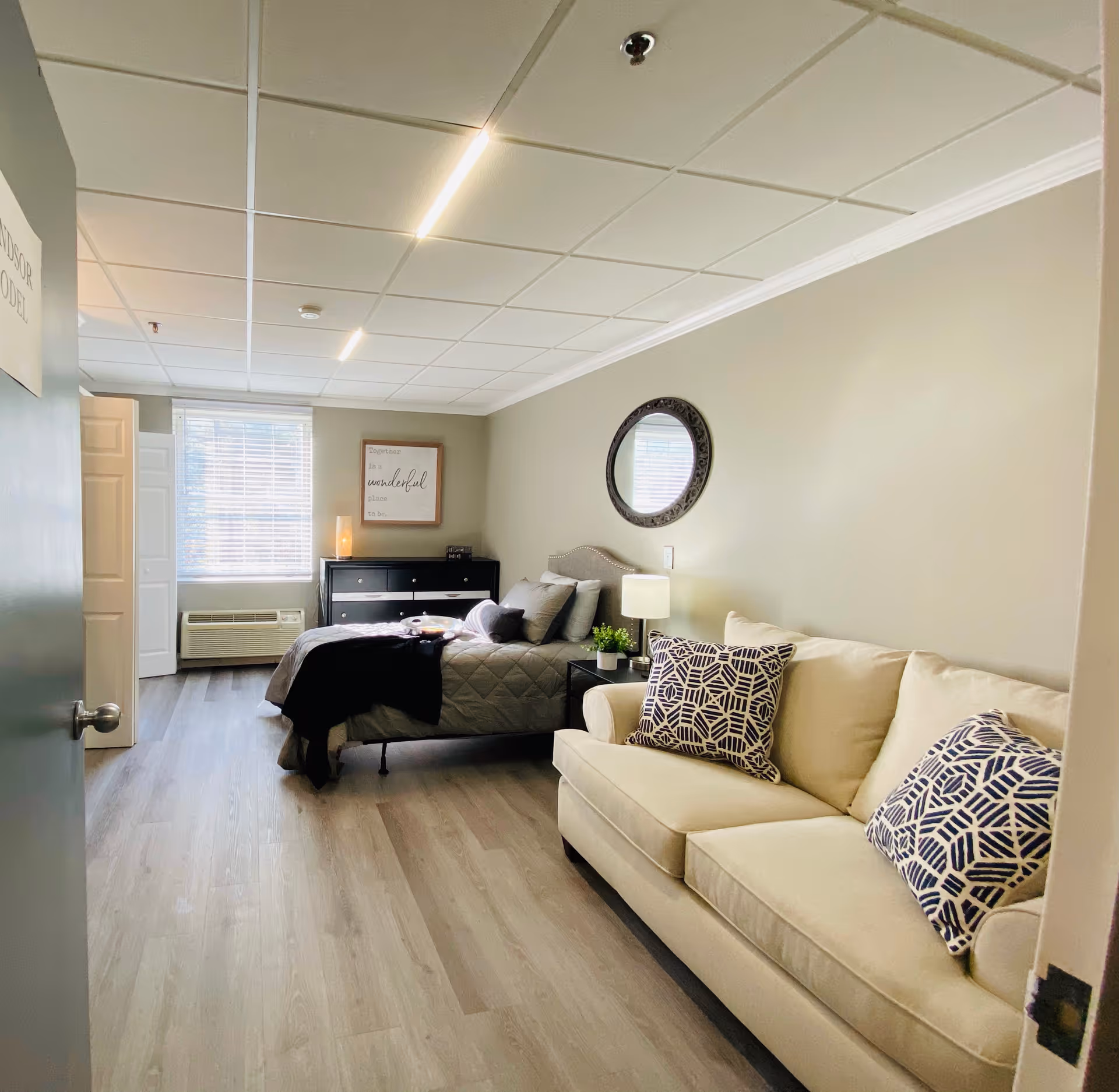 A bright and cozy senior living bedroom featuring a beige couch with patterned pillows on the right, a bed with gray bedding and multiple pillows in the center, a black dresser with a lamp and decor against the far wall, a round mirror above a bedside table with a lamp, and a window with blinds letting in natural light.