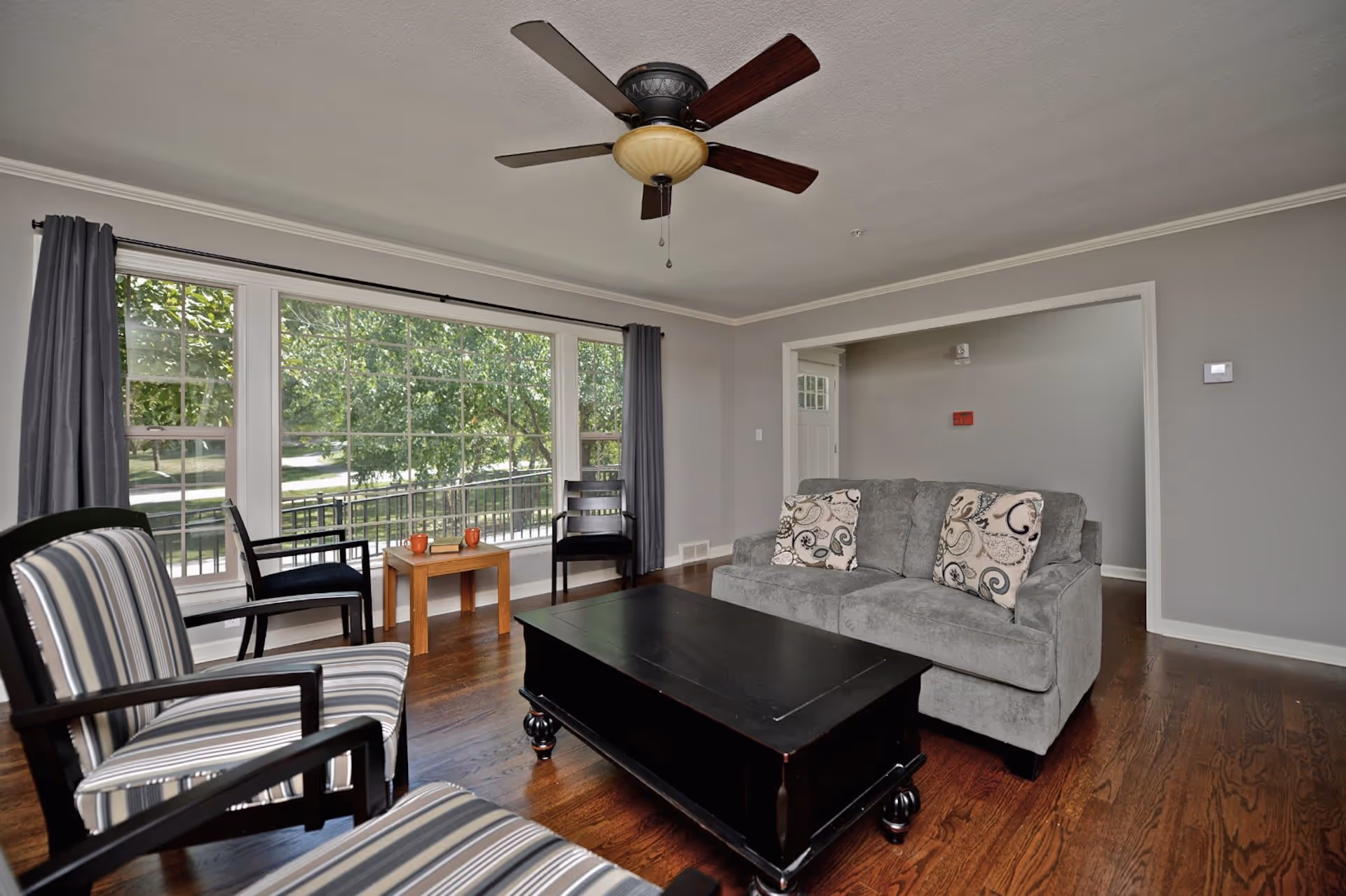 A cozy living room with a gray couch adorned with patterned pillows, two striped armchairs, a black coffee table, and a small wooden side table with two orange mugs and a book. Large windows with gray curtains allow natural light to brighten the room, and a ceiling fan with wooden blades hangs from the ceiling. The room has wooden flooring and light gray walls.