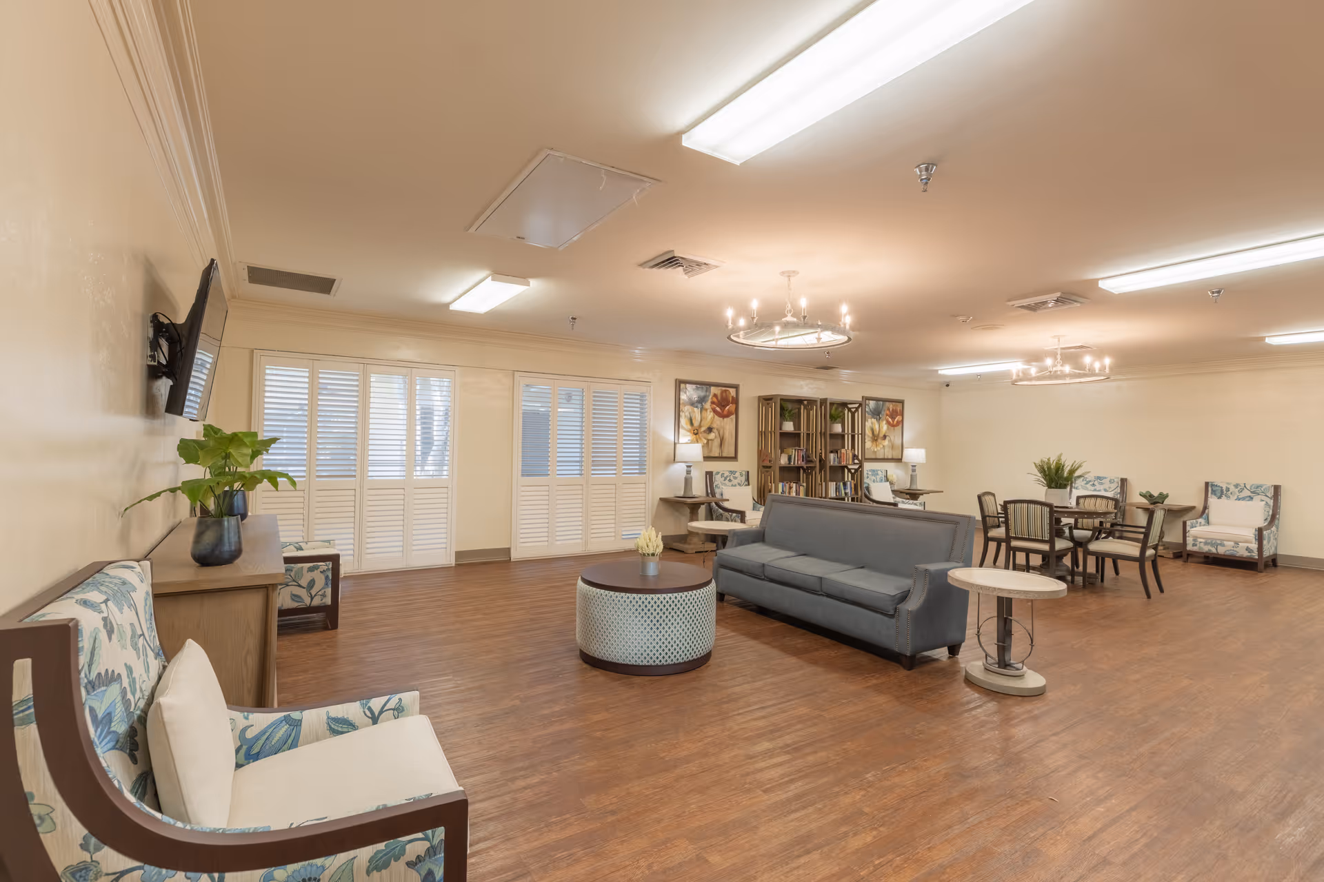 A spacious senior living facility common area with wooden flooring, beige walls, and ceiling lights. The room features a gray sofa, patterned armchairs, a round coffee table, side tables, a wall-mounted TV, a bookshelf with decorative items, and a dining table with chairs. Large windows with white shutters allow natural light into the room.