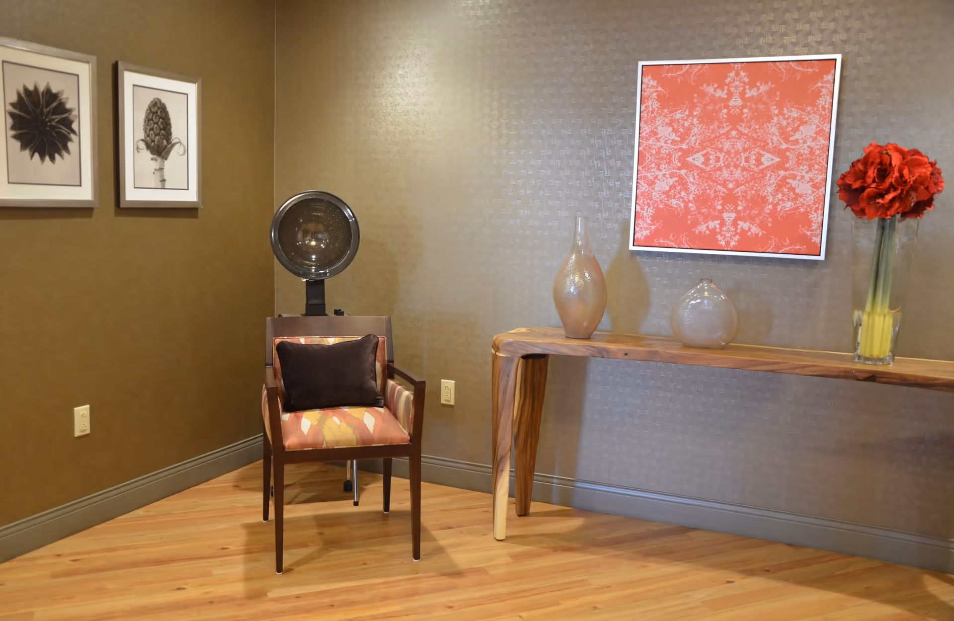 A corner of a room with a wooden floor and textured beige walls. There is a wooden chair with a patterned cushion and a dark pillow, positioned in front of a vintage hair dryer. On the right side, a wooden console table holds two decorative vases and a glass vase with red flowers. Above the table hangs a framed red and white abstract artwork. Two framed botanical prints are on the left wall.