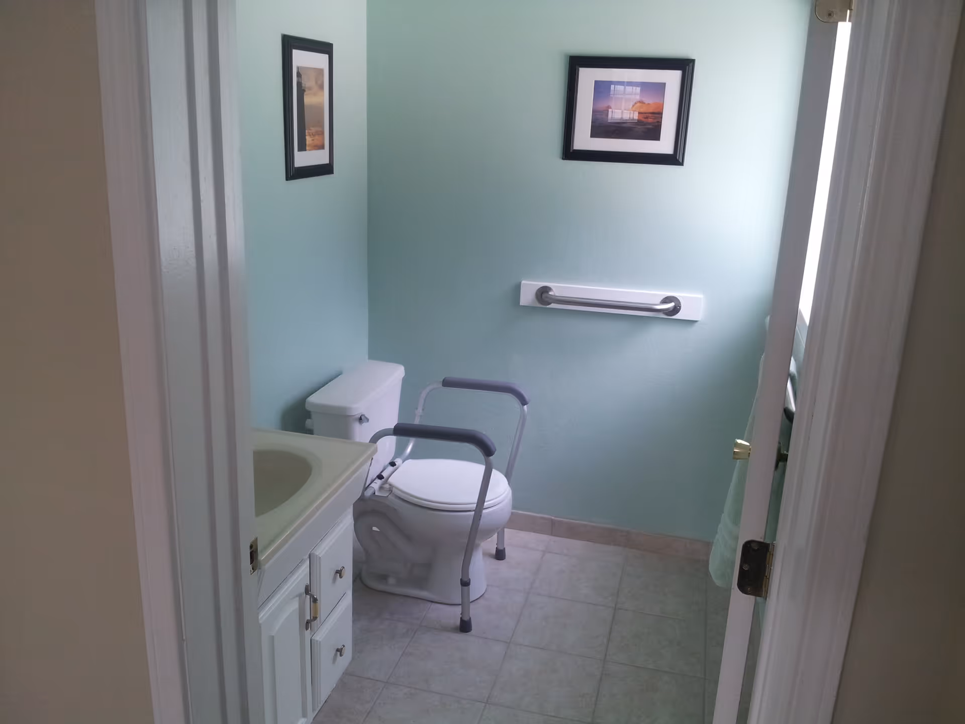 A small bathroom with light green walls, a white toilet equipped with safety rails, a white sink cabinet, a grab bar mounted on the wall, and two framed pictures hanging above the toilet.