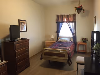 Simple assisted-living bedroom with a single hospital-style bed, dresser and TV, a window with curtains, and small side tables.