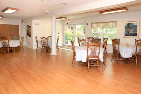 Bright dining room with round tables, white cloths, wooden chairs, and large windows.
